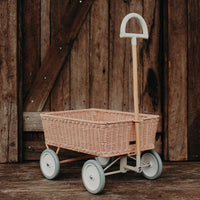 Olli Ella Rattan Wonder Wagon in rose with a woven basket wooden handle and pale wheels displayed against a rustic wooden backdrop