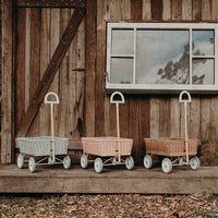 Three Olli Ella rattan wonder wagons in white pink and natural tan lined up outside a rustic wooden barn with vintage windows and woodland ground cover