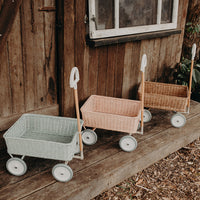 Three Olli Ella rattan Wonder Wagons in mint pink and natural lined up on a wooden porch outdoors with wheels and pull handles visible