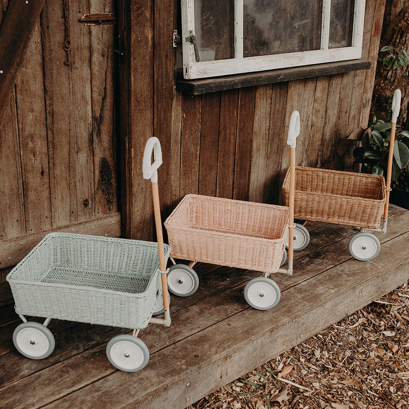 Three Olli Ella rattan Wonder Wagons in mint pink and natural lined up on a wooden porch outdoors with wheels and pull handles visible