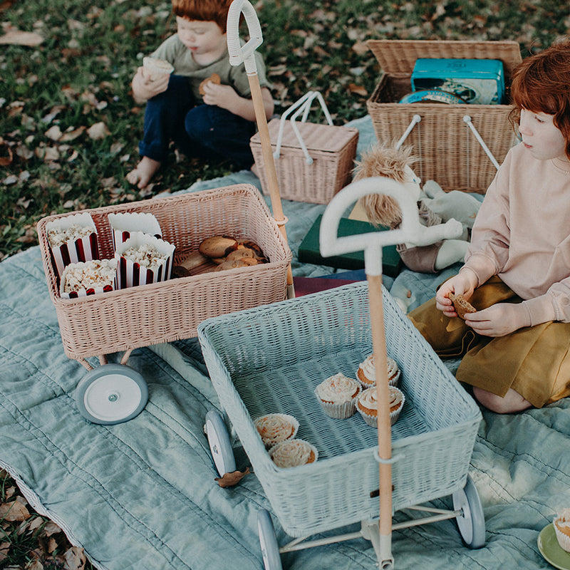 Children playing with a pastel rattan Wonder Wagon and woven baskets during an outdoor picnic with dolls and pretend food on a quilt by Olli Ella Kids Rattan