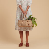 Olli Ella Lifestyle rattan basket with bamboo handles held by a person in a floral dress and filled with leafy greens against a warm neutral background
