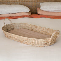 Olli Ella Nursery woven rattan baby changing basket with curved handles and a quilted cushioned liner resting on a softly styled nursery bed