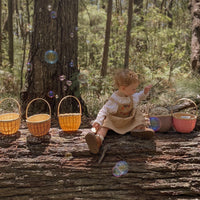 Child playing in a woodland beside colourful woven bamboo baskets including a berry pink Blossom Basket from Olli Ella Kids Rattan with bubbles floating nearby