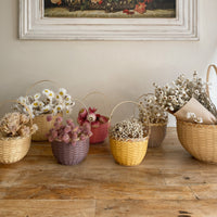 Small berry-colored woven basket with a curved handle holding dried flowers on a wooden table alongside other baskets in a warm home setting