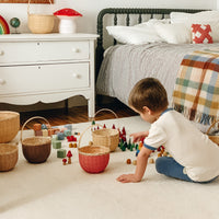 Child playing with colourful toys beside a small berry red bamboo basket in a cosy bedroom play scene.