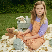Child playing outdoors with a vintage blue mini rattan bag beside a teddy bear and basket while sitting on a floral blanket with Olli Ella accessories nearby
