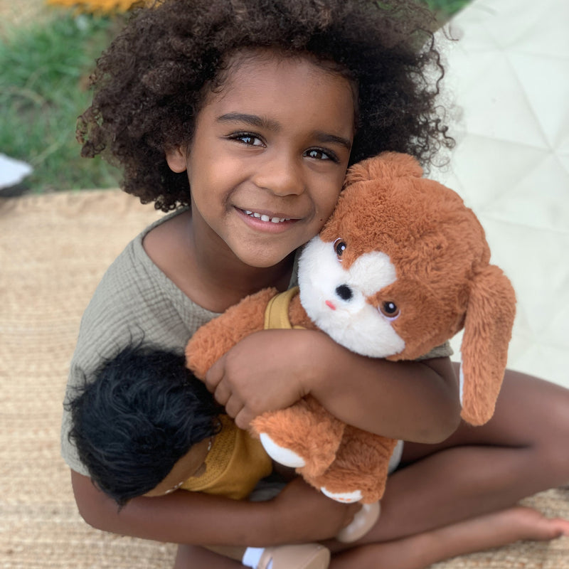 A smiling child cuddles a soft brown and white plush dog alongside a black haired doll indoors. Dinkum Pets Buddy inspires imaginative play.