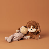 Soft brown plush Dinkum Dog cuddling a sleepy Dinkum Doll on a warm terracotta backdrop. A playful companion for imaginative play from Dinkum Pets