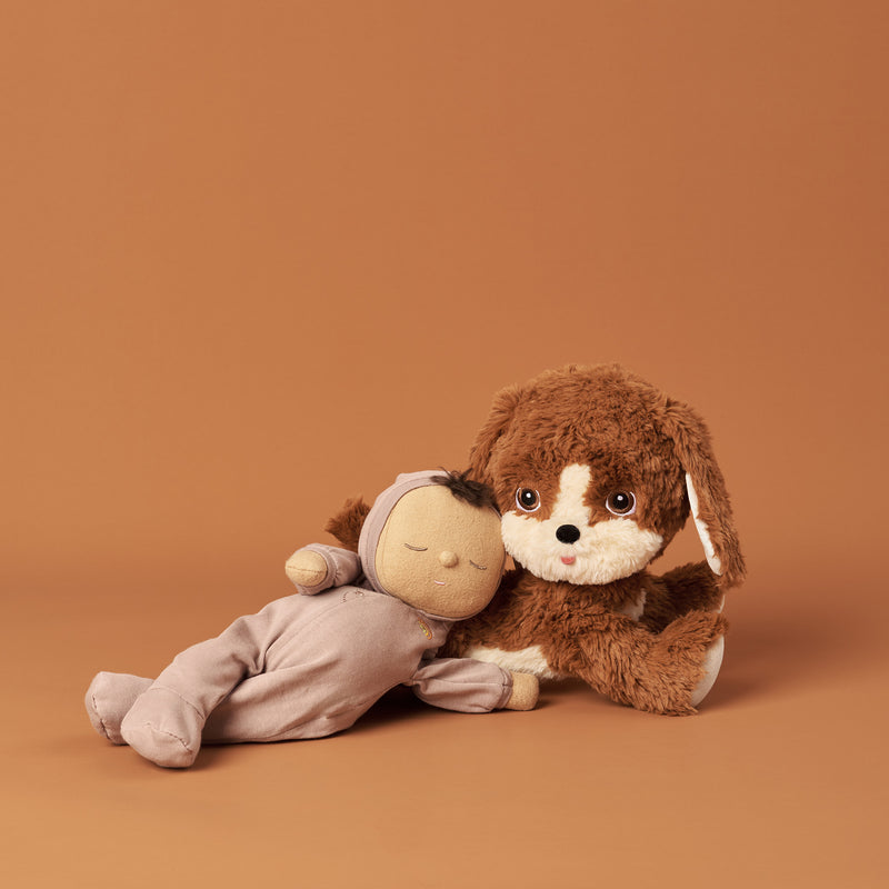 Soft brown plush Dinkum Dog cuddling a sleepy Dinkum Doll on a warm terracotta backdrop. A playful companion for imaginative play from Dinkum Pets