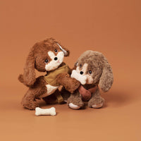 Two soft plush Dinkum Pets dogs play together with a white toy bone on a warm brown background. One wears a tan coat and the other a brown scarf.