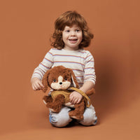 A smiling child cuddles a soft brown Dinkum Dog Buddy plush toy while seated against a warm terracotta background in this playful kids portrait
