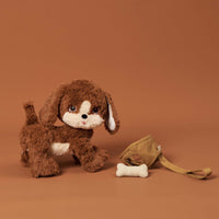 Brown plush Dinkum Pets puppy with a white face beside a tan dog harness and soft white bone on a warm brown backdrop for imaginative play