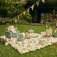 Outdoor garden play scene with an Olli Ella Kids Rattan chalk luggy alongside wicker baskets and soft dolls on a floral picnic blanket