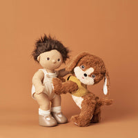 Dinkum Pets Buddy plush dog wearing a yellow harness stands beside a smiling Dinkum Doll in a warm studio setting for imaginative play