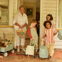 Children and an adult stand outside with wheeled rattan baskets including a pale chalk Luggy filled with treasures from Olli Ella Kids Rattan
