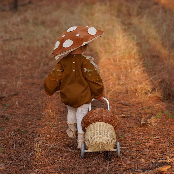 Dinkum Doll wearing a mushroom hat and brown outfit pulls a natural rattan Mushroom Luggy through a woodland setting from Olli Ella Kids Rattan