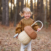Smiling child outdoors in a woodland carrying a natural two tone rattan mushroom luggy with a white handle from Olli Ella
