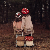 Two children wearing mushroom hats pull natural rattan mushroom luggys through a woodland setting for imaginative outdoor play by Olli Ella