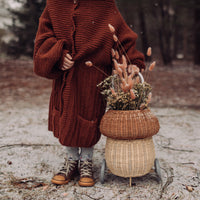Child in a cosy red knit coat pulls an Olli Ella Kids Rattan mushroom luggy filled with dried flowers through a frosty woodland setting