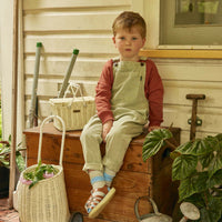 Child sitting on a wooden chest beside a chalk coloured rattan Luggy basket filled with flowers in a warm outdoor setting by Olli Ella Kids Rattan