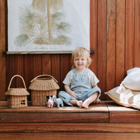 Child sitting beside a handmade rattan house basket with a sturdy handle and small dolls on a wooden bench in a warm nursery setting by Olli Ella Kids Rattan
