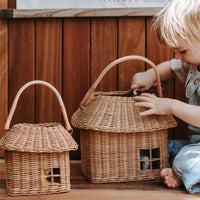 Child playing with a large handmade rattan house basket beside a smaller matching basket with handles and window openings from Olli Ella Kids Rattan