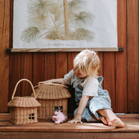 Child playing with a handmade rattan house basket and smaller basket beside a plush toy on a wooden bench from Olli Ella Kids Rattan