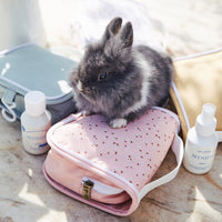 Olli Ella Kids pink floral wash bag with a fluffy black bunny and travel toiletries on a sunny beach
