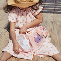 Child in a floral dress opening a pink See Ya wash bag with a mesh pocket and playful purple character detail during a sunny outdoor adventure